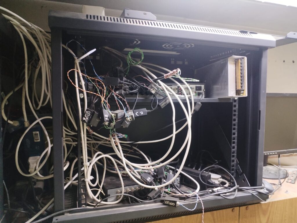La imagen muestra un **gabinete de CCTV en estado desordenado antes de ser reparado**. En su interior se encuentran varios **DVRs (grabadores de video digital)**, junto con una gran cantidad de cables desorganizados y conexiones inadecuadas. Algunos de los problemas visibles incluyen: 🔴 **Cables de alimentación y video enredados y sin gestión**, lo que dificulta el mantenimiento y la identificación de fallos. 🔴 **Mala distribución de los dispositivos**, lo que puede generar sobrecalentamiento y reducir la vida útil del equipo. 🔴 **Conexiones expuestas y sin protección**, aumentando el riesgo de cortocircuitos y pérdida de señal en las cámaras. 🔴 **Ausencia de etiquetado en los cables**, dificultando la localización de cada conexión y afectando la operatividad del sistema. 📌 **Un gabinete de CCTV en este estado puede generar fallos en la transmisión de video, pérdida de grabaciones y fallos en el monitoreo de seguridad.** Es fundamental una reorganización profesional para garantizar un funcionamiento óptimo y seguro.