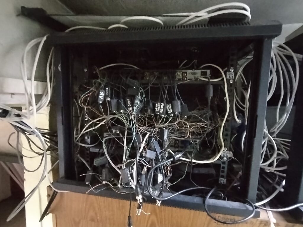 La imagen muestra un **gabinete de telecomunicaciones en estado crítico antes de ser reparado**. Se observa un **desorden extremo en el cableado**, con múltiples hilos y conexiones sin organización ni identificación adecuada.  

Algunos de los problemas visibles incluyen:  
🔴 **Cables enredados y sin gestión**, lo que dificulta la identificación y solución de fallas.  
🔴 **Conectores y dispositivos desordenados**, sin una distribución lógica dentro del gabinete.  
🔴 **Posible interferencia y fallos eléctricos**, debido a la falta de canalización y protección adecuada.  
🔴 **Riesgo de cortocircuitos**, ya que algunos cables parecen estar expuestos o mal conectados.  

📌 **Este tipo de instalaciones pueden causar fallos constantes en sistemas de seguridad, redes de datos y telecomunicaciones.** Un mantenimiento profesional es clave para evitar problemas y optimizar su funcionamiento.